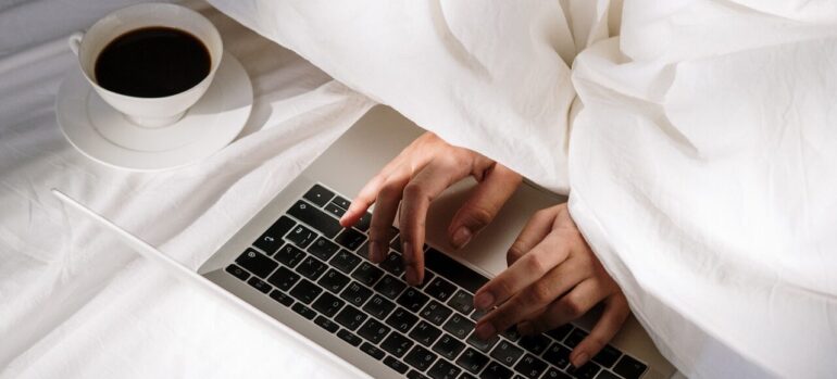 A person typing on a keyboard under the sheets next to a cup of coffee.