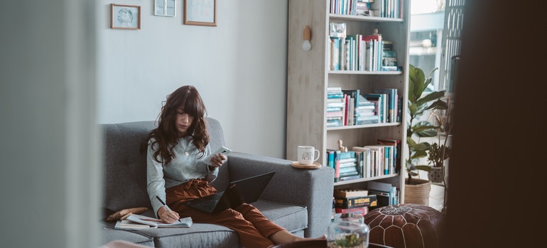 A woman using a notebook and her laptop on a couch.