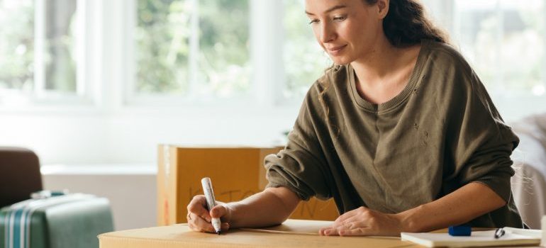 A woman in a brown sweater writing something on a box.