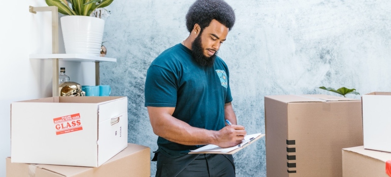 a man in a blue uniform making a list of items while standing in a room full of cardboard boxes to depict the evaluation of the moving from NYC to LA process