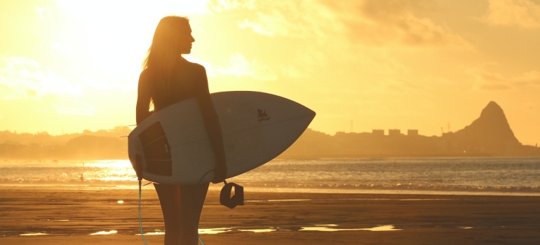 a woman holding a surfing board on the beach with the sunset on the horizon